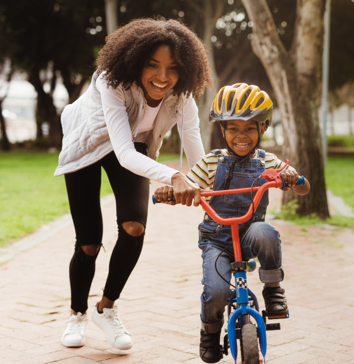 a mother teaching her son how to ride a bicycle outdoors. The mother and son are happy and smiling as she holds and guides his handle bars